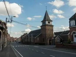 Plombier Oupeye devant l'église Notre-Dame d'Oupeye, rue pavée bordée d'arbres et maisons en briques typiques.